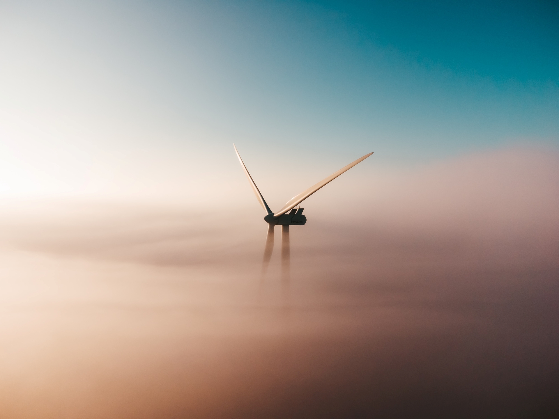 Wind turbine blades emerging above fog under a clear blue sky at sunrise or sunset.