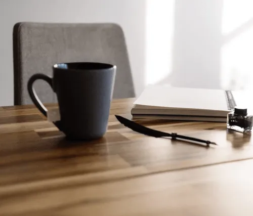 Black coffee mug, feather quill pen, ink bottle, and a blank notebook on a wooden table with a gray chair in the background.