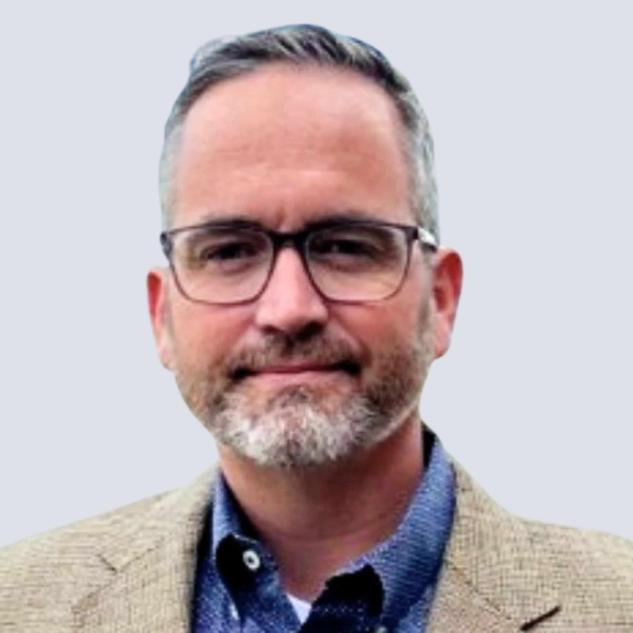 Middle-aged man with short salt-and-pepper hair, beard, and glasses wearing a beige blazer and blue shirt outdoors with mountains blurred in the background.