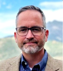 Middle-aged man with short salt-and-pepper hair, beard, and glasses wearing a beige blazer and blue shirt outdoors with mountains blurred in the background.