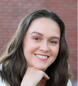 Smiling woman with long dark hair resting her chin on her hand against a blurred brick wall background.