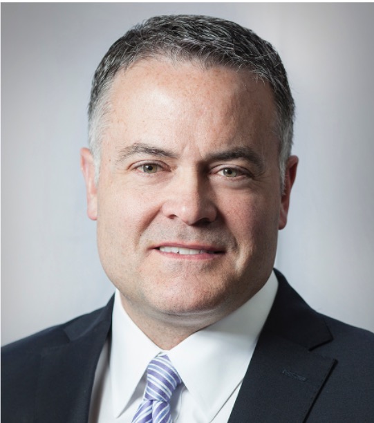 Professional headshot of a middle-aged man wearing a dark suit, white shirt, and striped tie, smiling slightly against a plain light background.