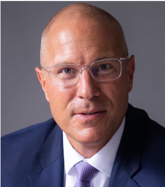 Middle-aged man with glasses wearing a dark suit and white shirt against a plain gray background.