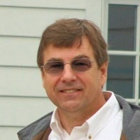 Middle-aged man with brown hair wearing glasses, a white shirt, and a gray jacket, smiling slightly outdoors.