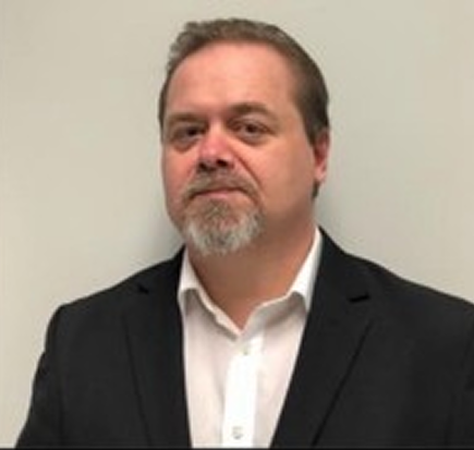 Portrait of a man with short hair and a goatee wearing a white shirt and black blazer against a plain light background.