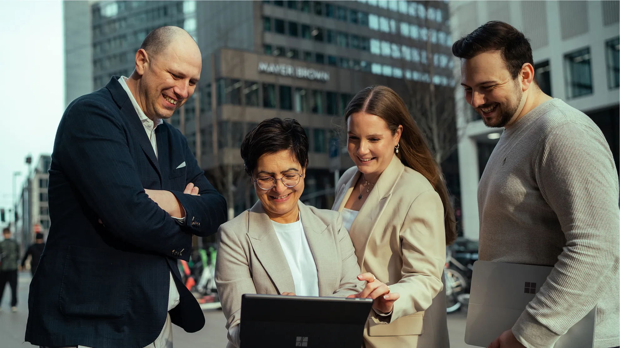 Vier Geschäftsleute, zwei Frauen und zwei Männer, schauen lächelnd auf ein Tablet im urbanen Umfeld.