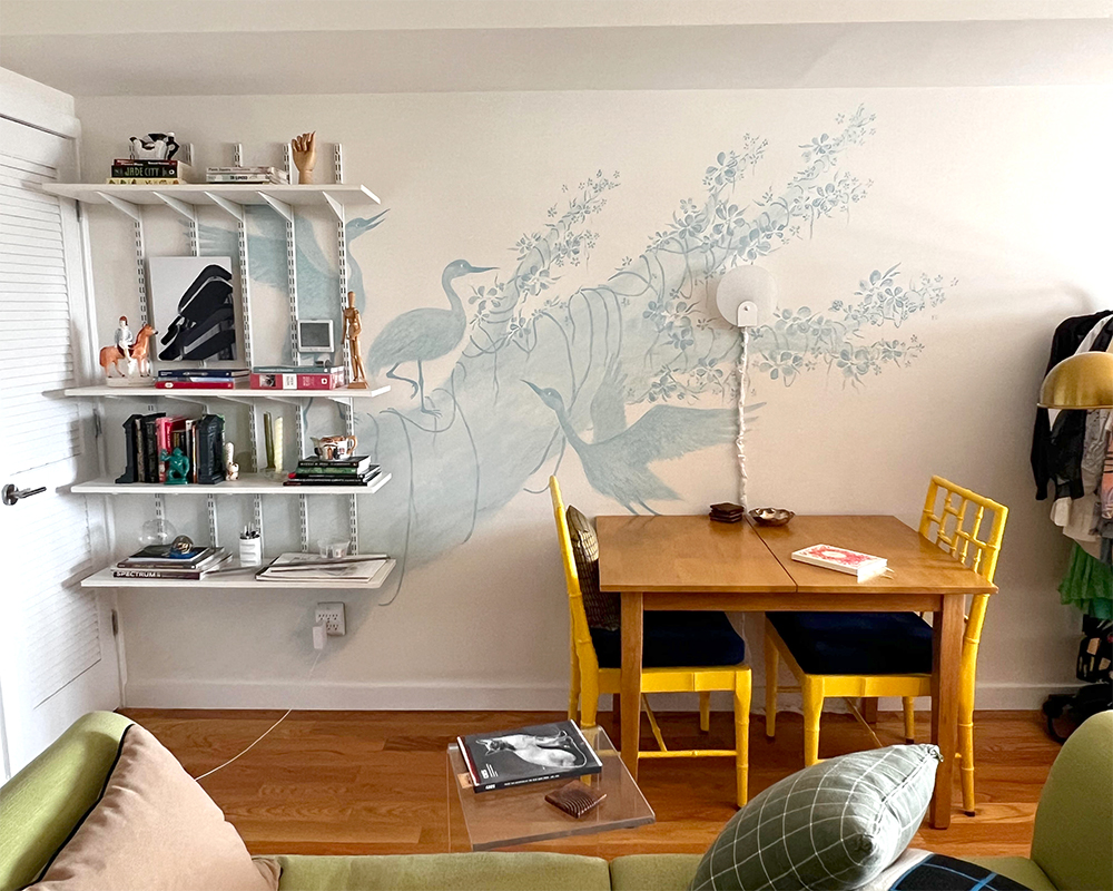 The table where I work. The image shows an apartment with a desk and a bookshelf against a wall. The wall has a mural on it depicted cranes on a tree.