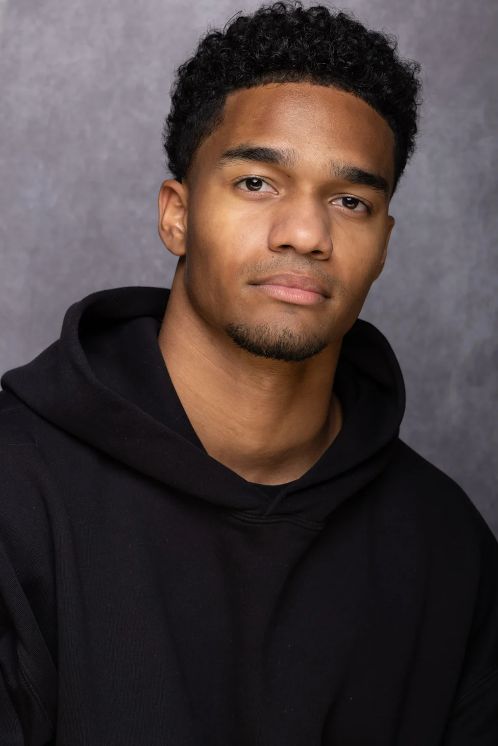 Young man with curly hair wearing a black hoodie against a gray backdrop.
