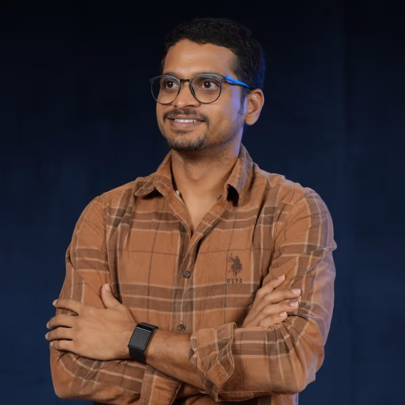Man with glasses wearing a brown plaid shirt with crossed arms smiling against a dark background.