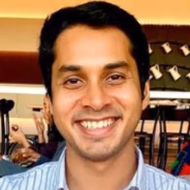 Smiling man with short black hair wearing a blue striped shirt, indoors with blurred background.