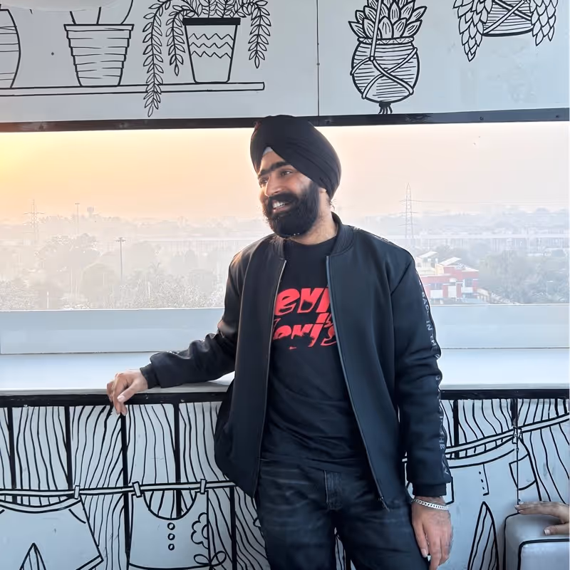 Man wearing a black turban and jacket smiling while leaning on a counter with plant-themed wall art behind him.