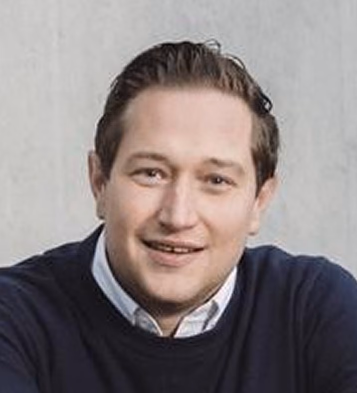Portret van een man met lichtbruine haren, gekamd naar achteren, die glimlacht en een wit overhemd onder een donkerblauwe trui draagt tegen een grijze achtergrond.