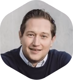 Portret van een glimlachende man met bruin haar, gekleed in een donkerblauwe trui en een lichtblauw overhemd, voor een grijze achtergrond.
