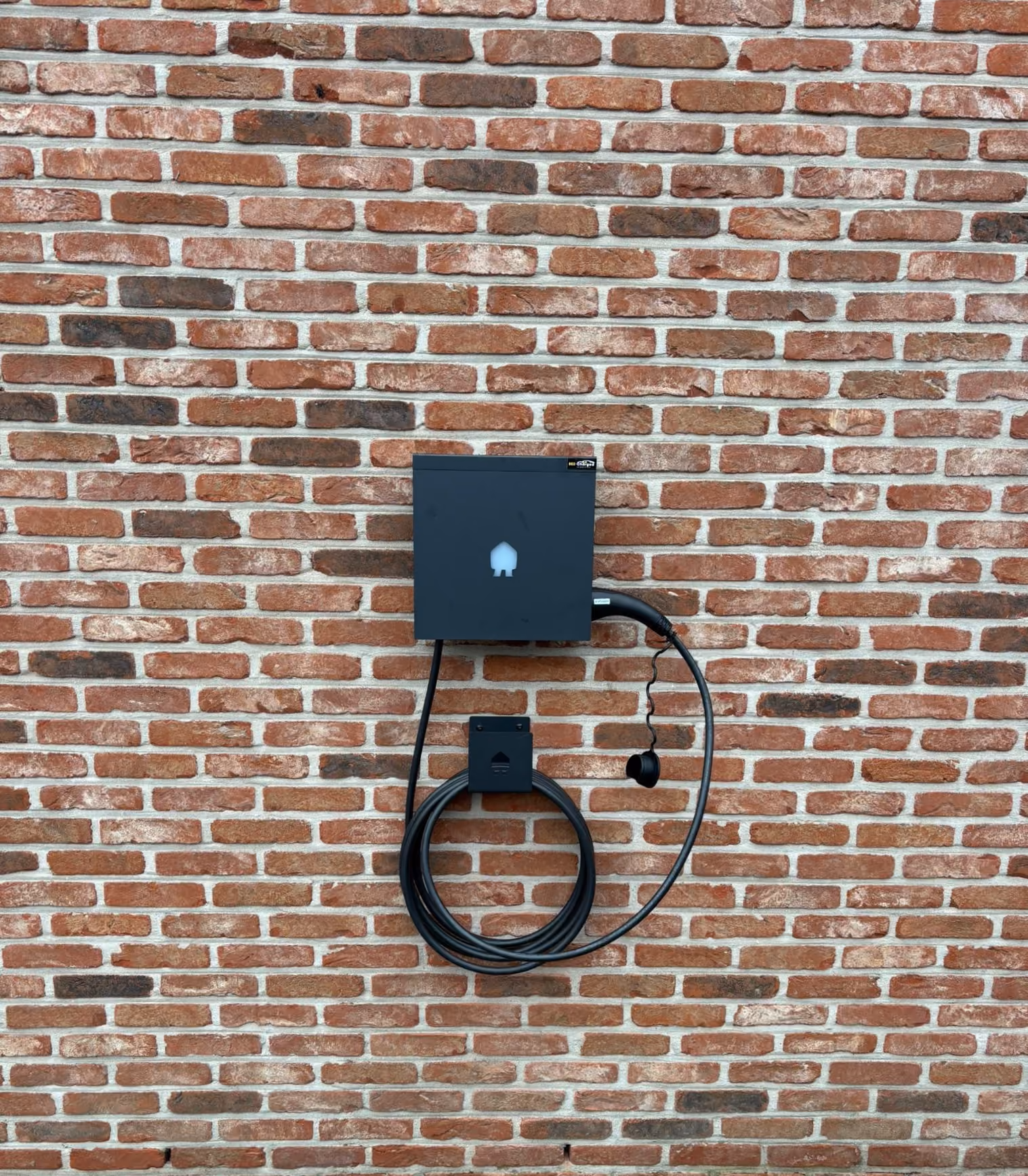 Zwarte elektrisch oplaadstation met oprolbare kabel gemonteerd aan een bakstenen muur.