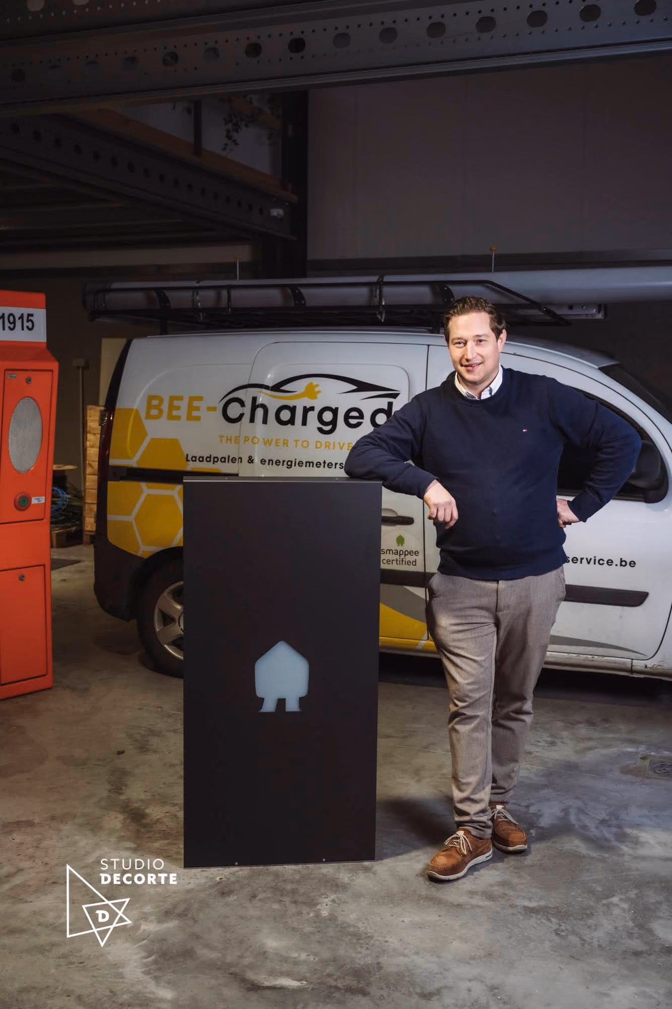 Man in a navy sweater and beige pants leans on a tall black charging station in a garage, with a white van behind him branded with Bee-Charged logo and text about charging stations and energy meters.