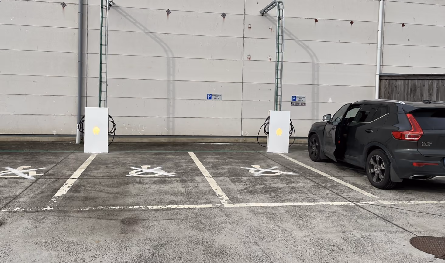 Parkeerplaatsen voor elektrische voertuigen met oplaadpunten bevestigd aan een grijze muur; een zwarte Volvo XC40 staat geparkeerd bij het meest rechtse oplaadpunt.