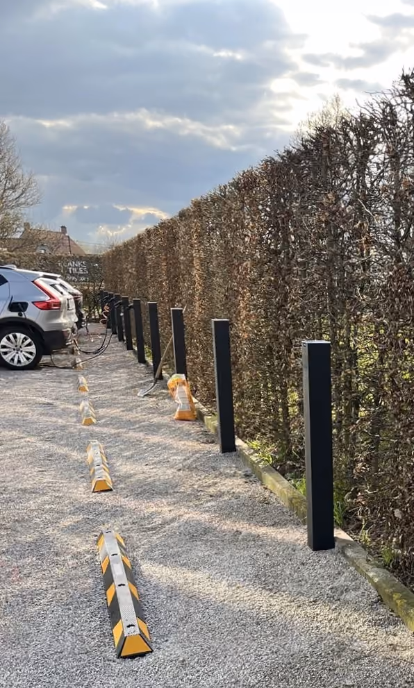 Parkeerplaats met elektrische auto's die aan het opladen zijn, omringd door hoge, nette hagen en meerdere parkeerblokken op grindbodem onder een bewolkte lucht.