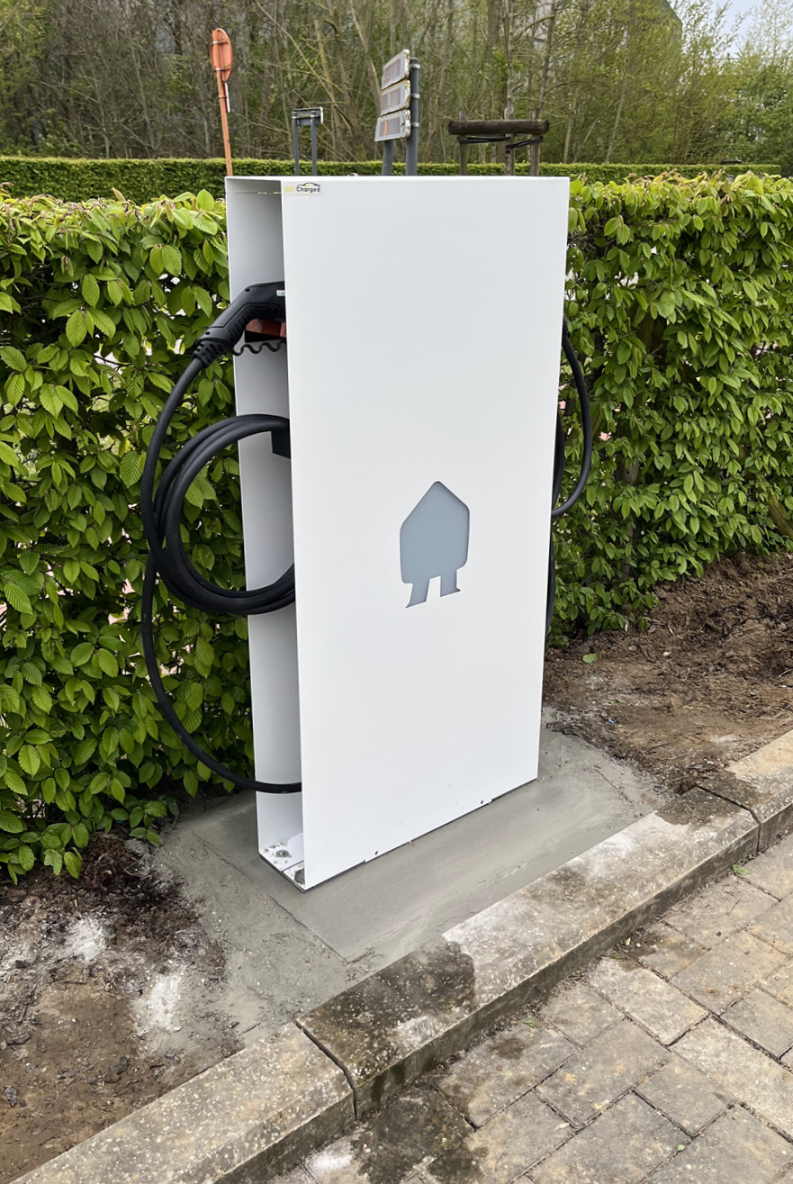 Witte elektrische laadpaal met een kabel en stekker, geplaatst op een betonnen ondergrond naast een groene haag, met straatstenen op de voorgrond.