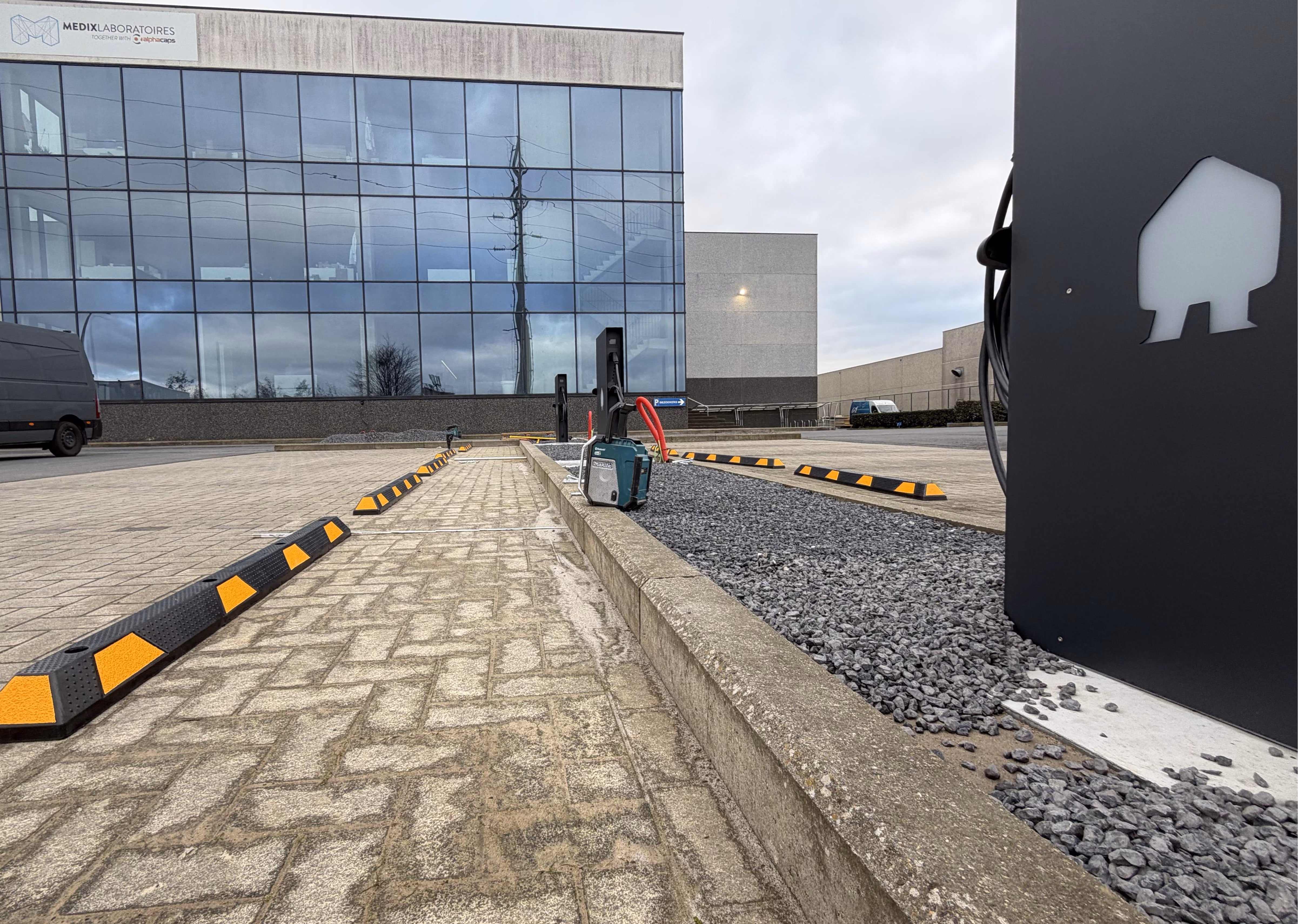 Parkeerplaatsen buiten een modern kantoorgebouw met grote ramen en het bord Medix Laboratoires aan de gevel, waarbij elektrische laadpalen zichtbaar zijn langs een strook met grijze grindstenen.