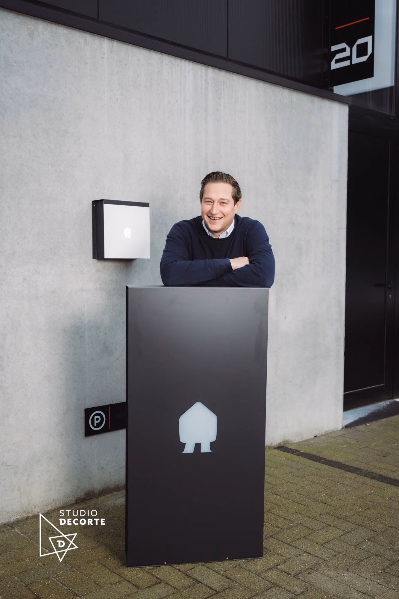 Een glimlachende man in een donkerblauwe trui leunt met zijn armen op een zwart bord met een wit huispictogram buiten voor een grijze betonnen muur.