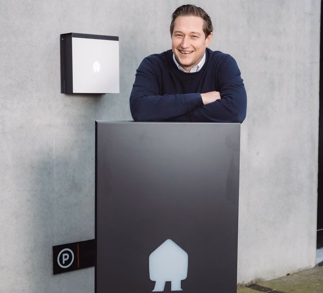 Een man in een donkerblauwe trui leunt lachend op een groot grijs object met een wit huisvormig symbool, tegen een lichte betonnen muur met een kleiner vergelijkbaar object aan de muur.