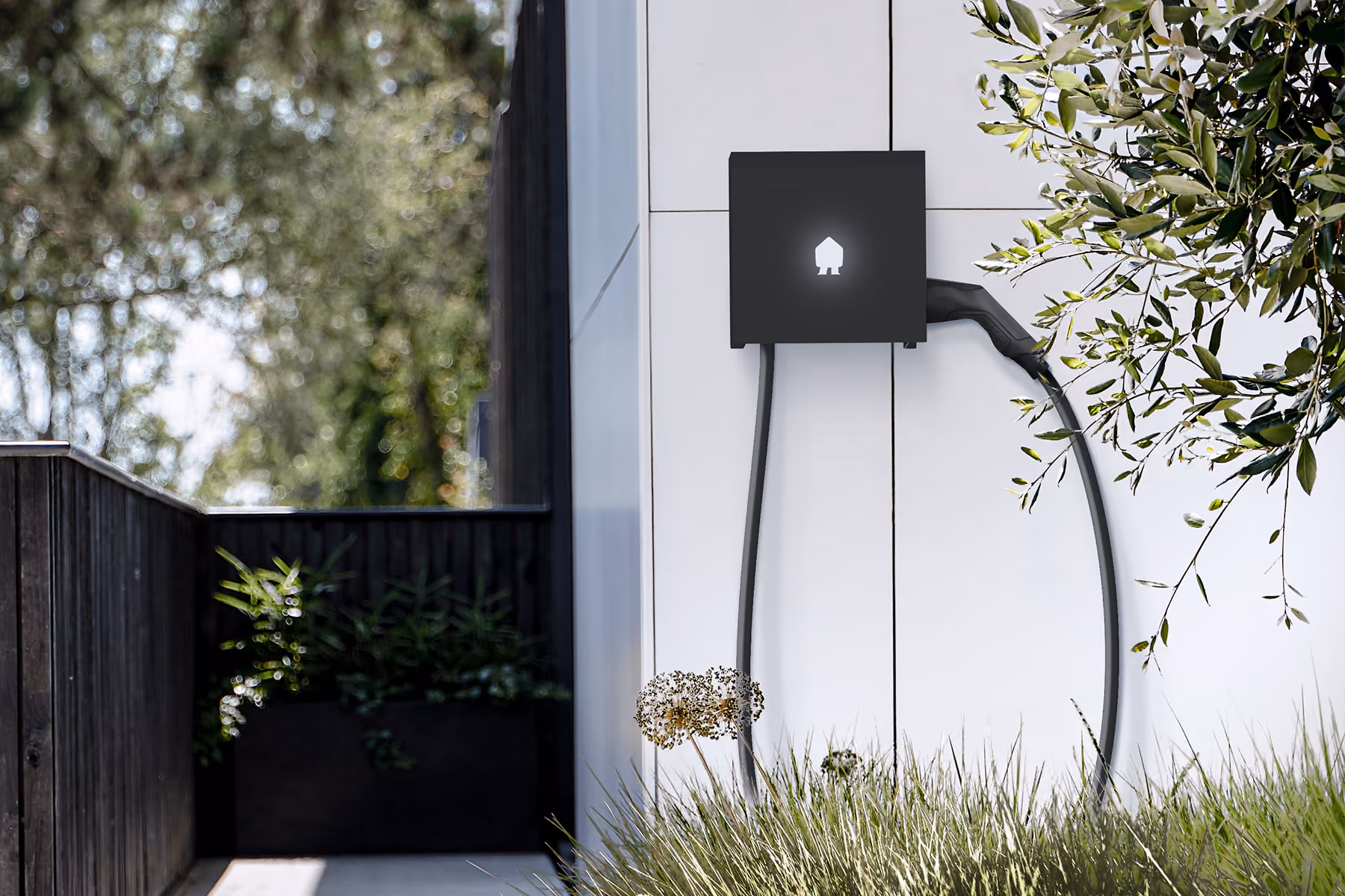 Elektrisch laadstation bevestigd aan een witte muur buiten, met een zwarte kabel en een verlicht Smappee logo, omgeven door groen gras en takken.