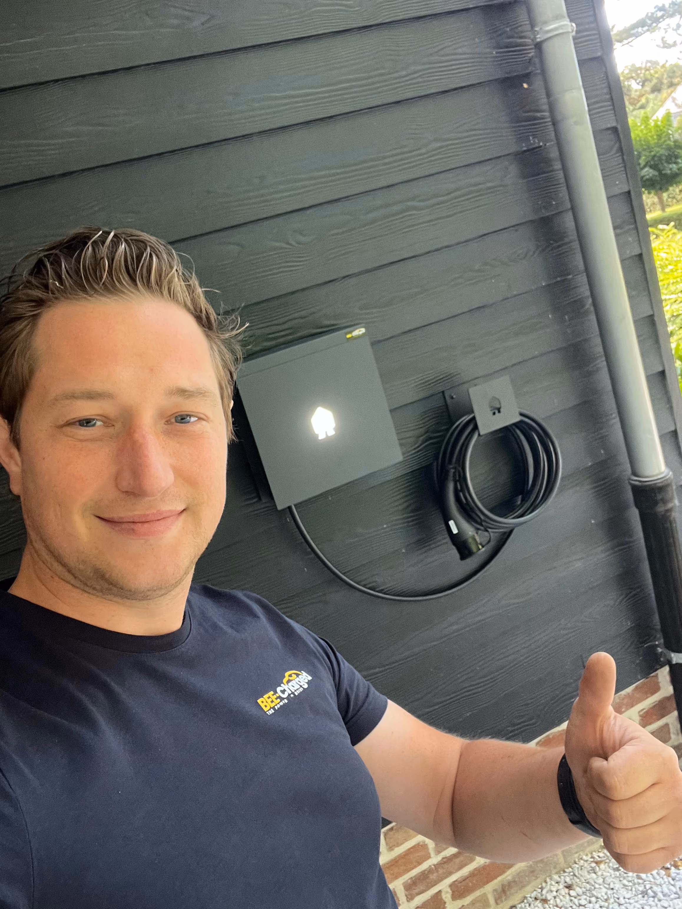Dries in een zwart Bee-Charged-shirt steekt zijn duim omhoog bij een zwarte muur met een gemonteerde laadpaal voor elektrische voertuigen.