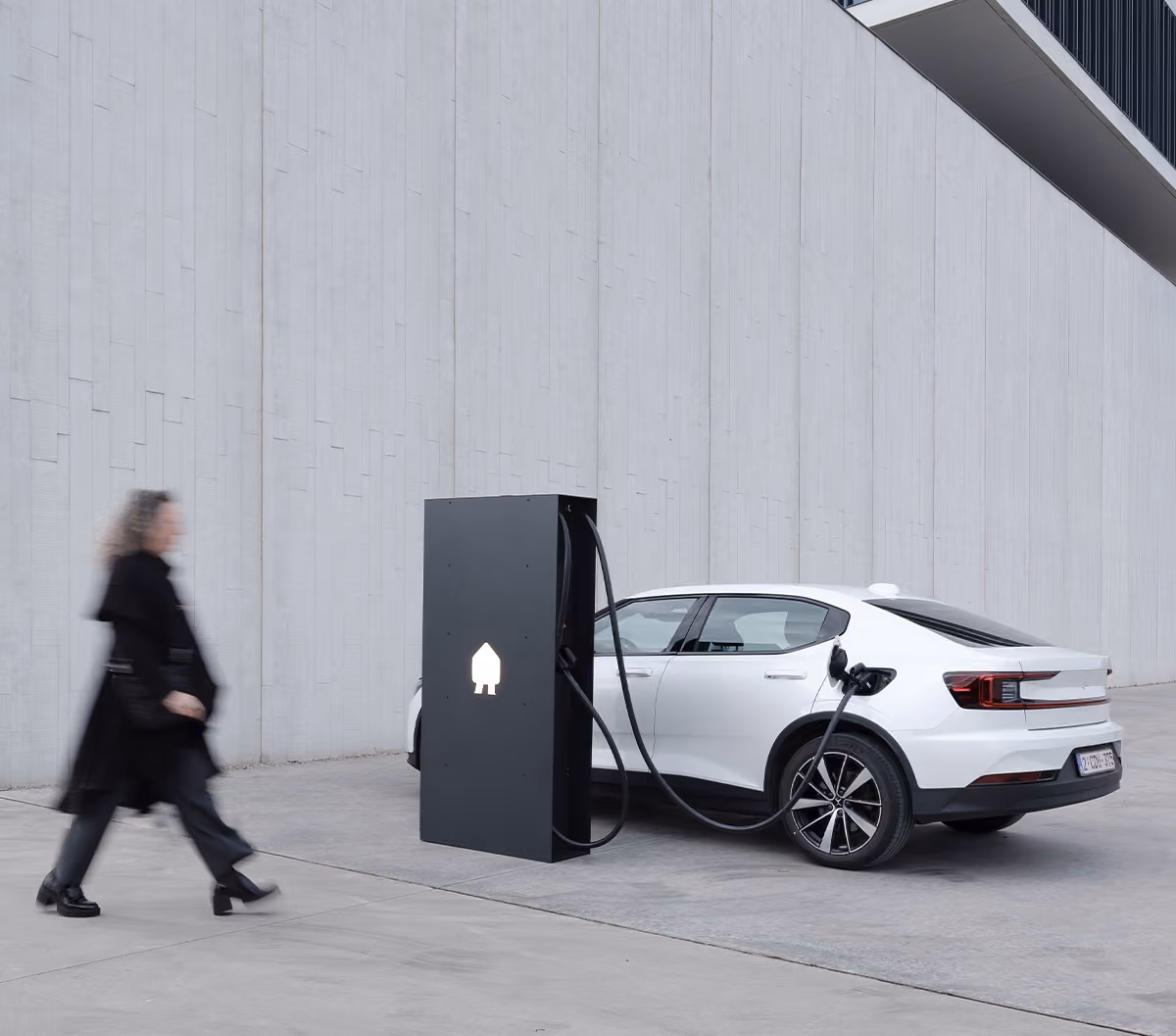Witte elektrische auto aangesloten op een zwarte laadpaal bij een betonnen gevel, met een persoon die langsloopt.