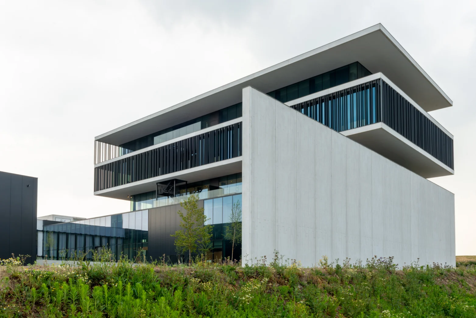 Een modern, minimalistisch gebouw met witte betonnen muren, grote glazen ramen, zwarte verticale lamellen en een plat dak, omgeven door groen.