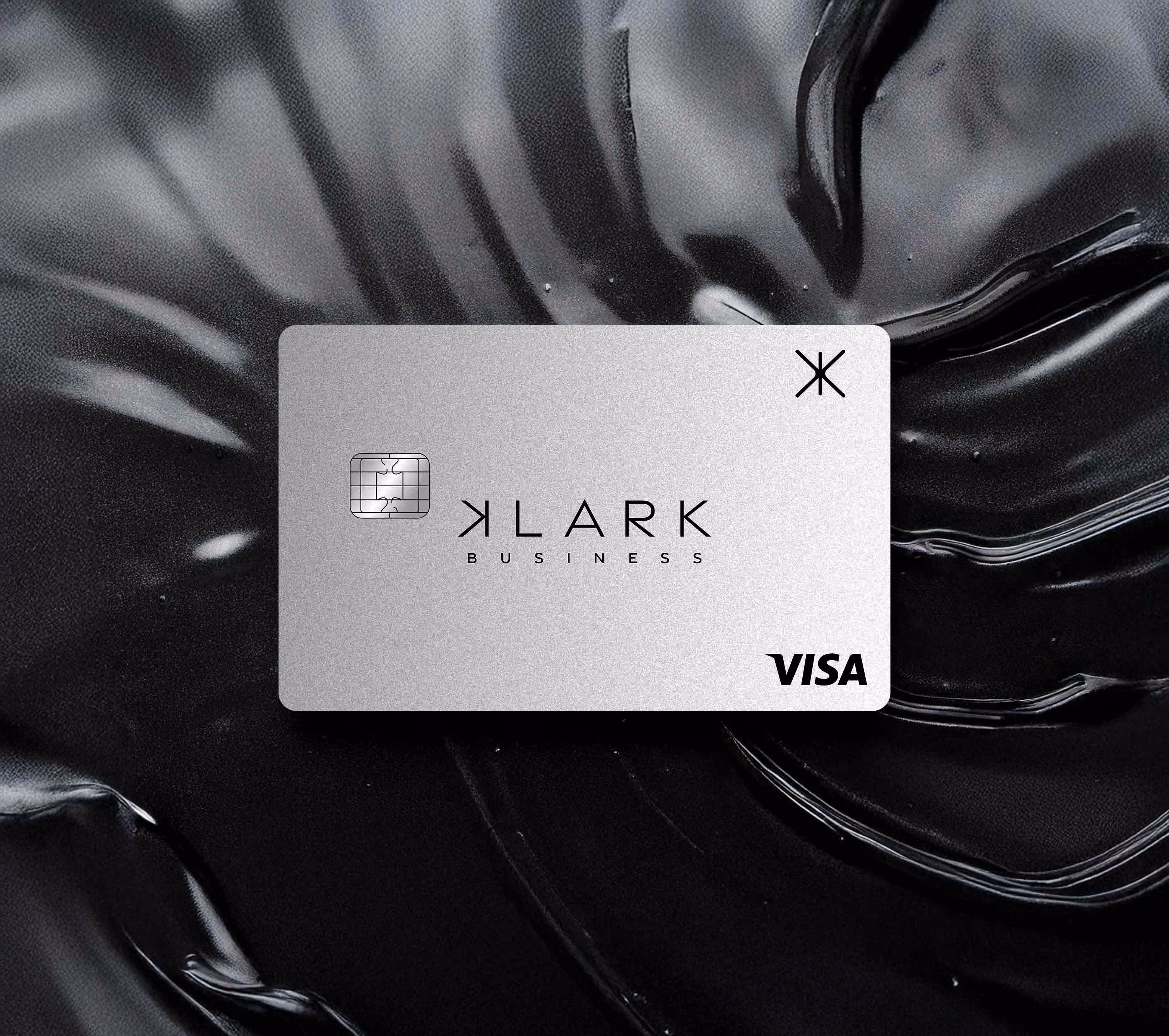 Carte bancaire grise Klark Business avec puce et logo Visa sur un fond noir texturé.