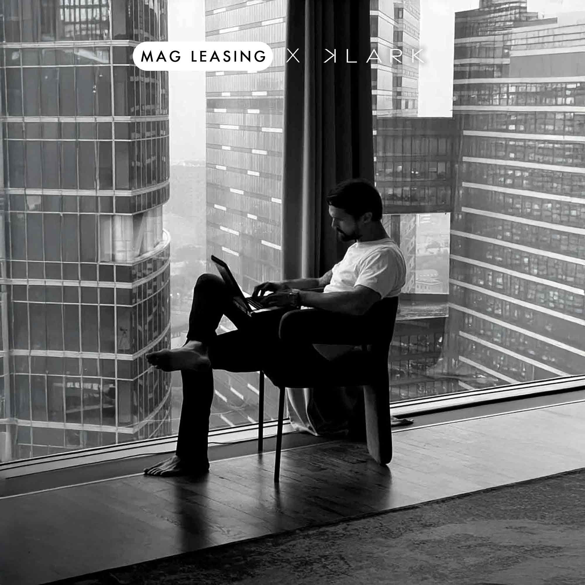 Homme assis pieds nus sur une chaise devant une large baie vitrée, travaillant sur un ordinateur portable avec vue sur des gratte-ciels en ville.