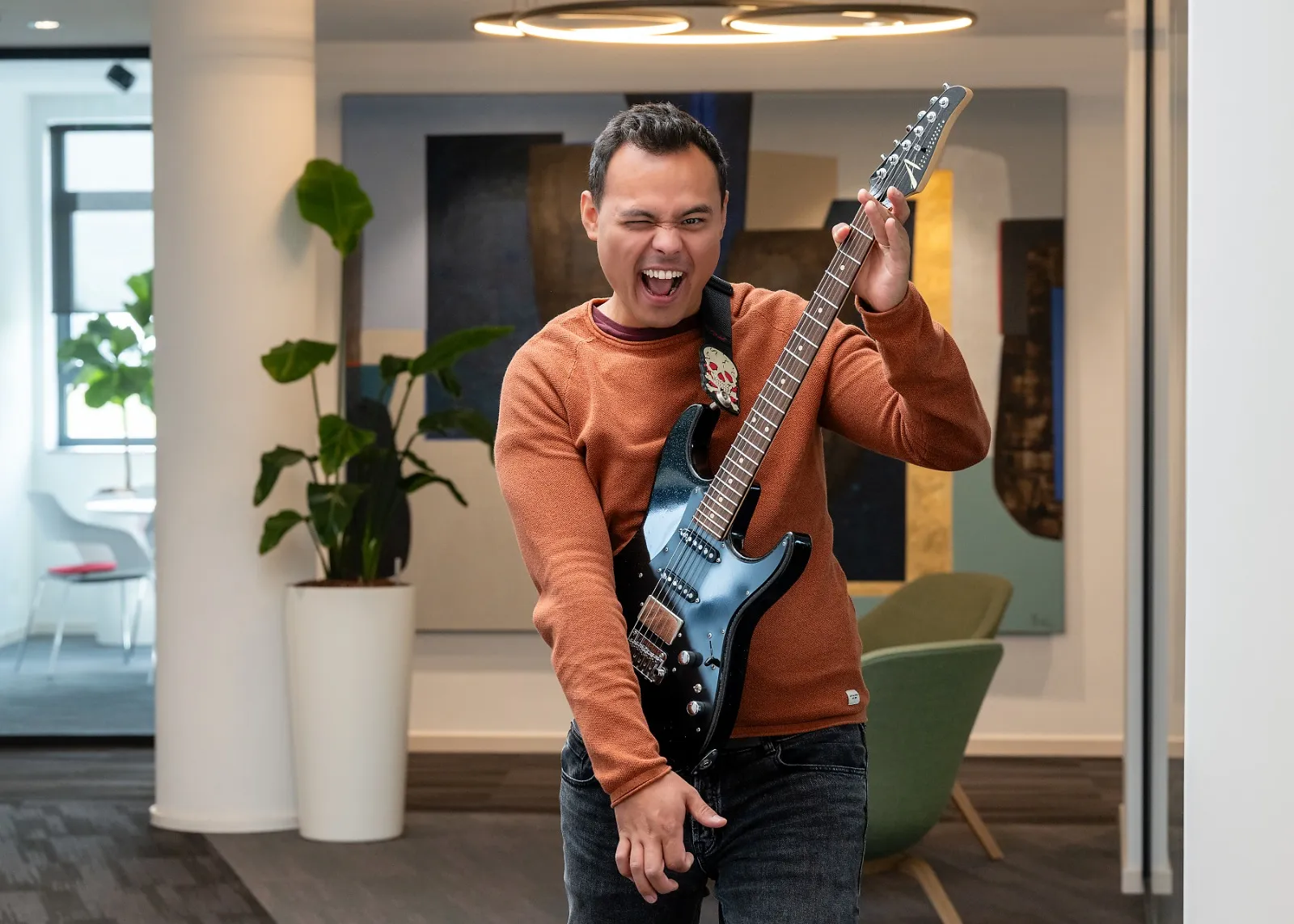 Kevin Struik en pull orangé portant une guitare électrique noire en signe de rock dans un intérieur moderne.