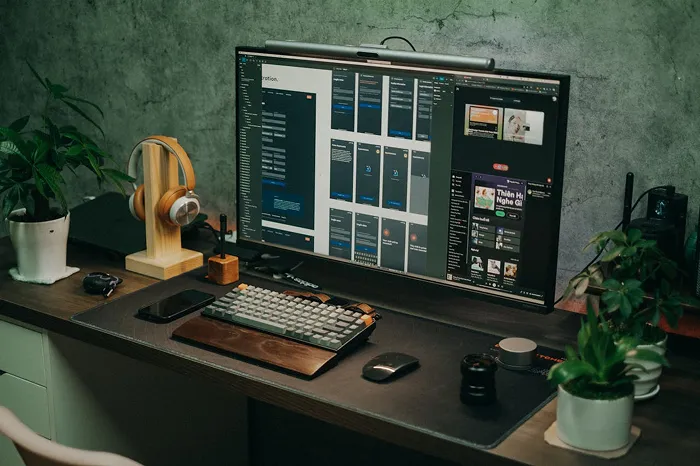 Bureau avec un grand écran ordinateur affichant un logiciel de conception, clavier mécanique, souris, casque sur support en bois, plusieurs plantes vertes sur le bureau.