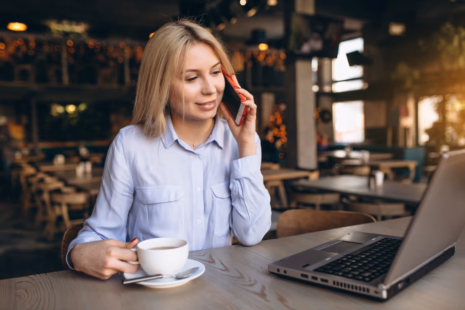 Sieviete ar zilu krekliņu sarunājas pa telefonu, sēžot kafejnīcā pie galda ar kafijas krūzi un klēpjdatoru.