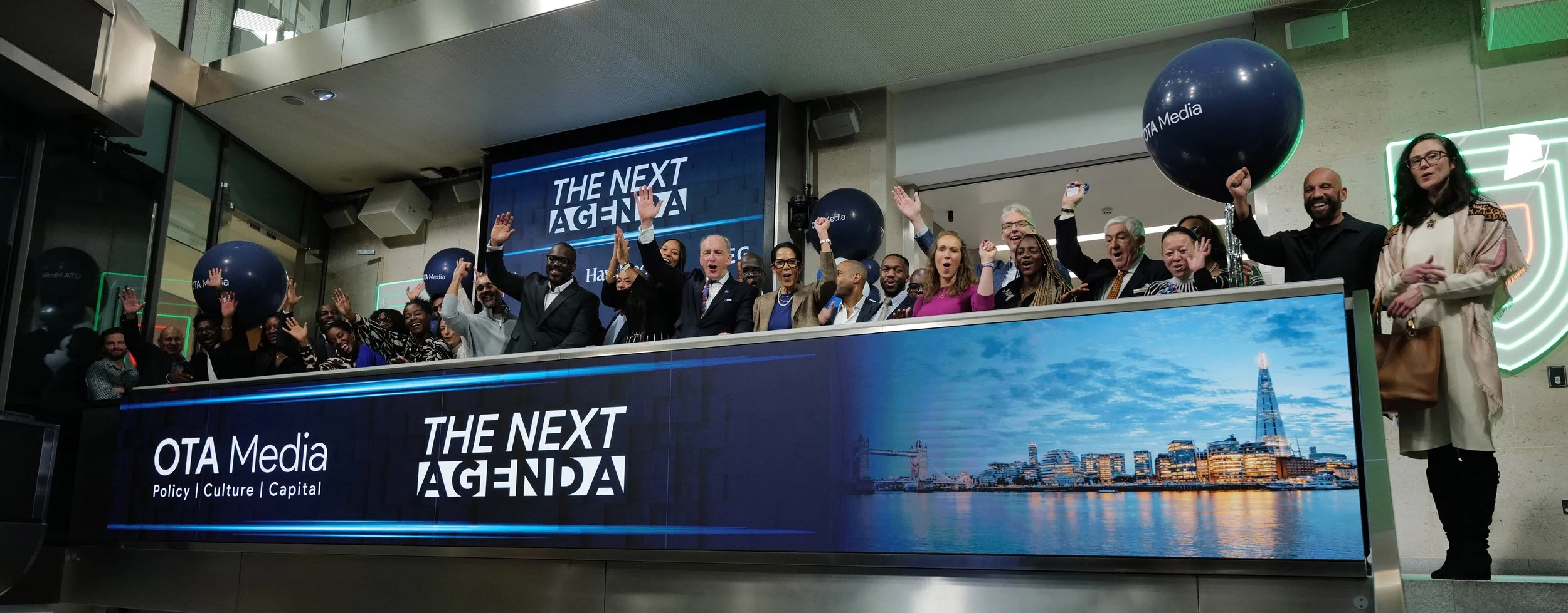 Group of diverse people standing behind a large digital display with OTA Media and The Next Agenda logos, celebrating indoors.