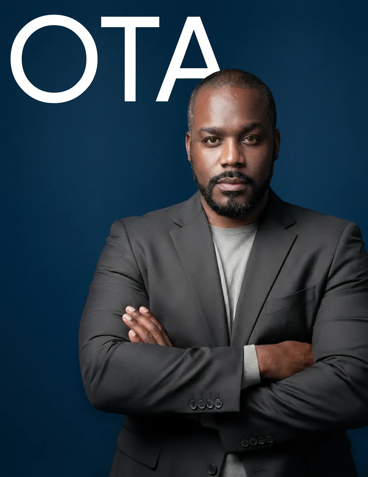Confident man with crossed arms wearing a dark blazer against a blue background with the text 'OTA'.