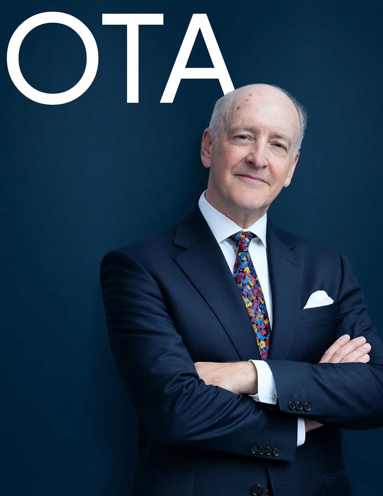 Confident older man in a dark suit with colorful tie and arms crossed against a dark blue background with the letters OTA.