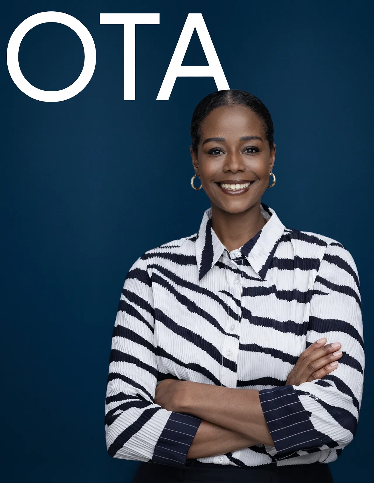Smiling woman with dark hair wearing gold hoop earrings and a white and black striped blouse, standing with arms crossed against a blue background.