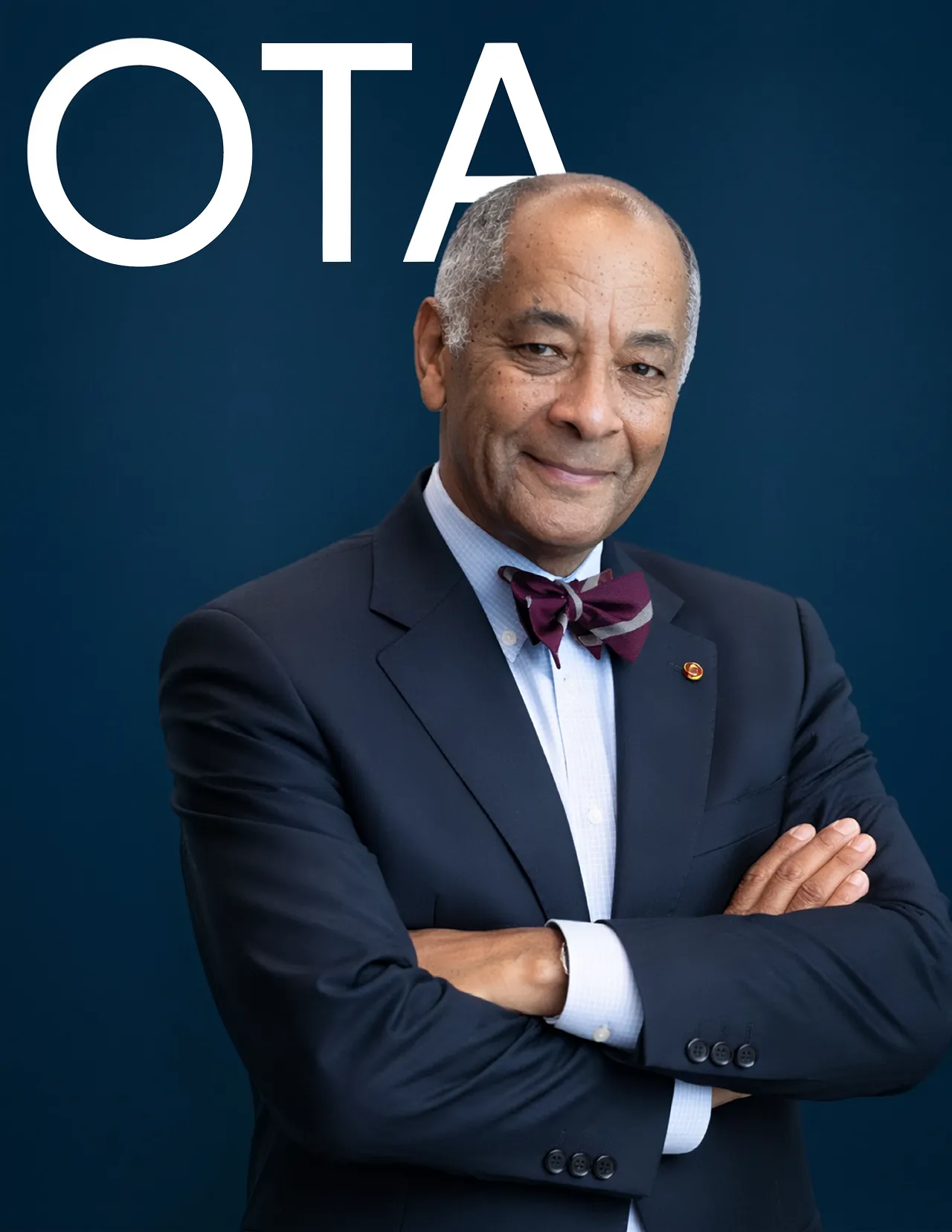 Smiling older man in a dark suit and burgundy bow tie standing with arms crossed against a dark blue background with partial white letters.