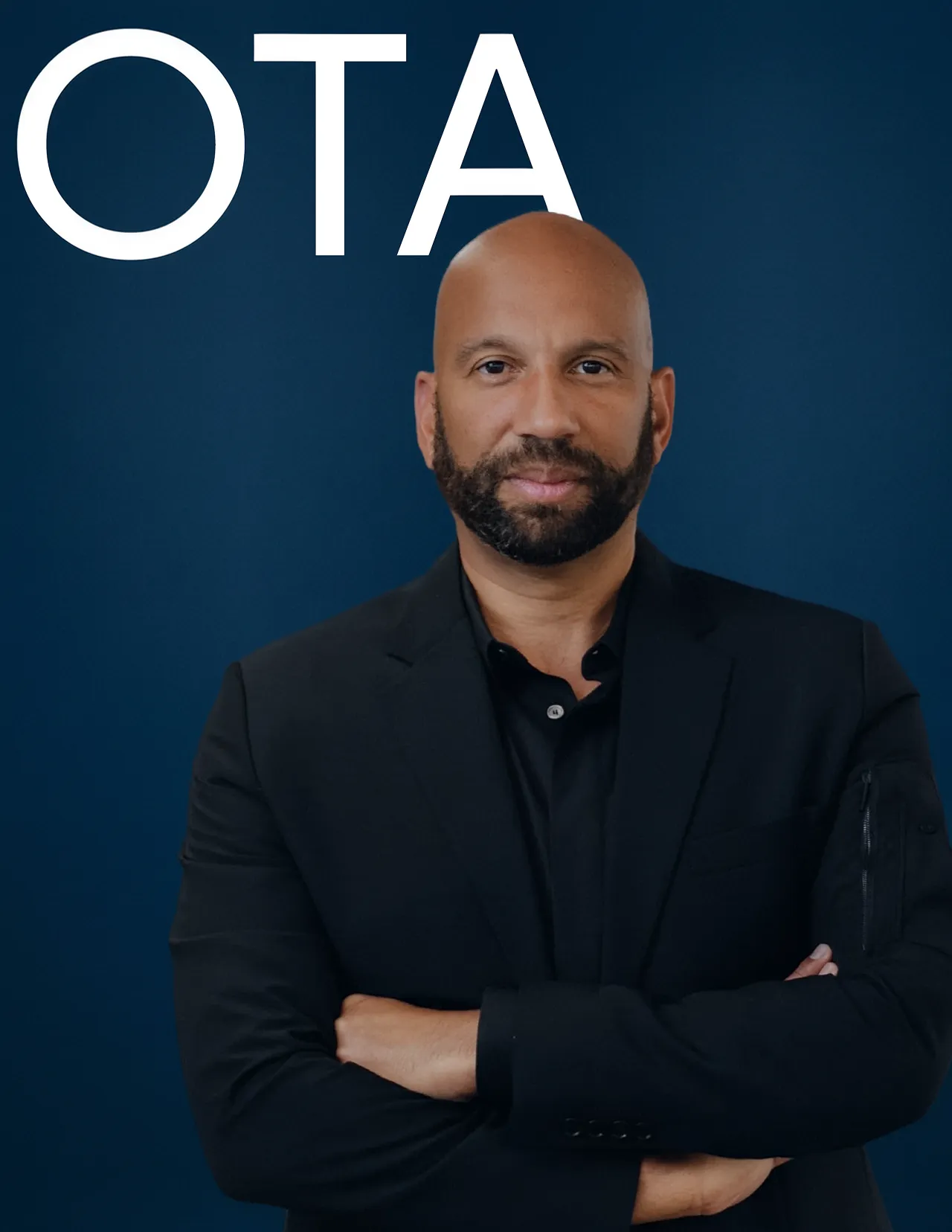 Bald man with a beard dressed in a black suit with arms crossed against a dark blue background with large white text 'OTA' above his head.