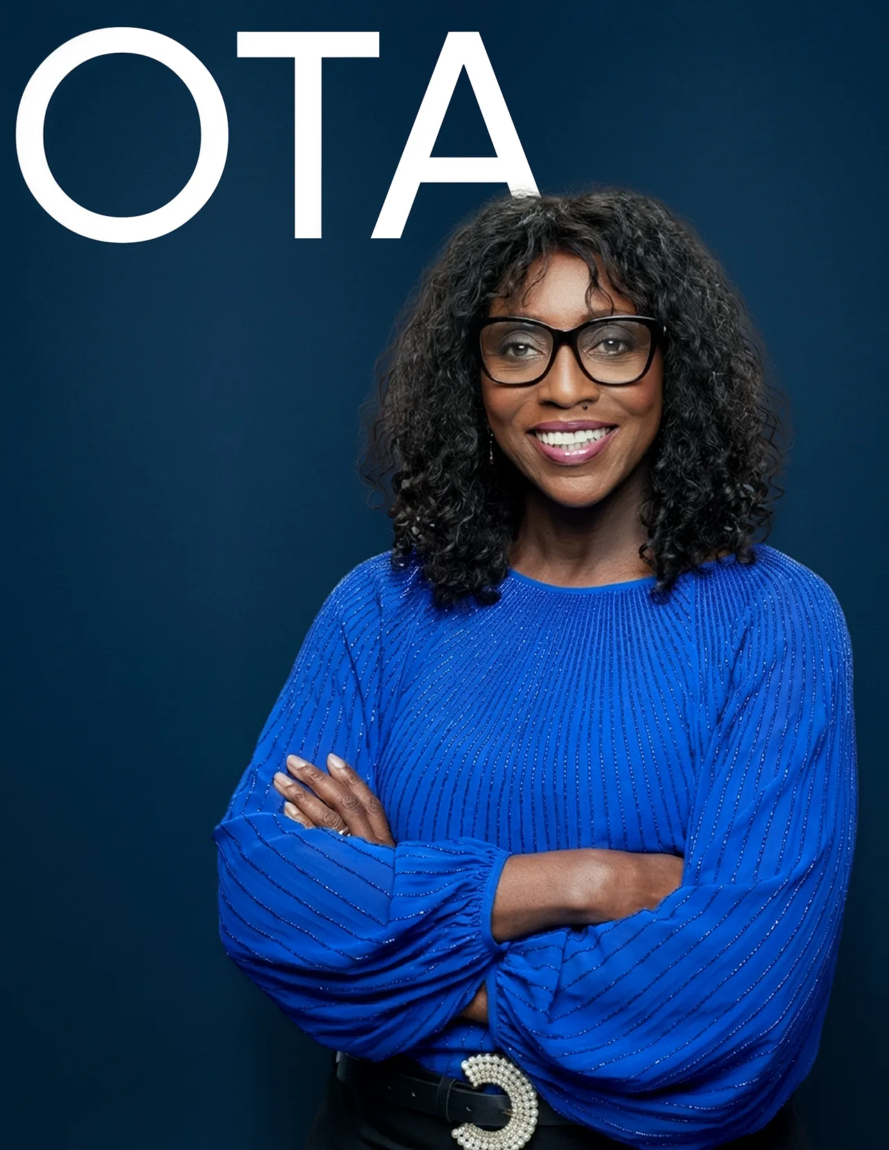 Smiling woman with curly hair and glasses wearing a blue blouse with crossed arms in front of a dark blue background with white letters OTA.