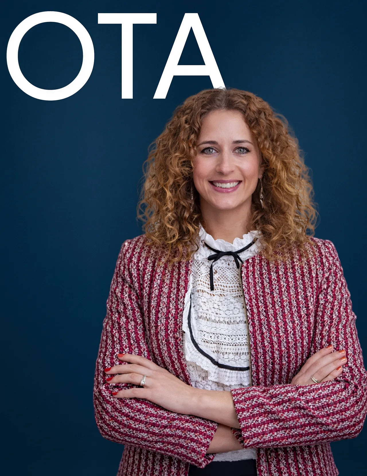 Smiling woman with curly hair wearing a red patterned blazer and white lace blouse standing with arms crossed against a dark blue background with large white letters 'OTA'.
