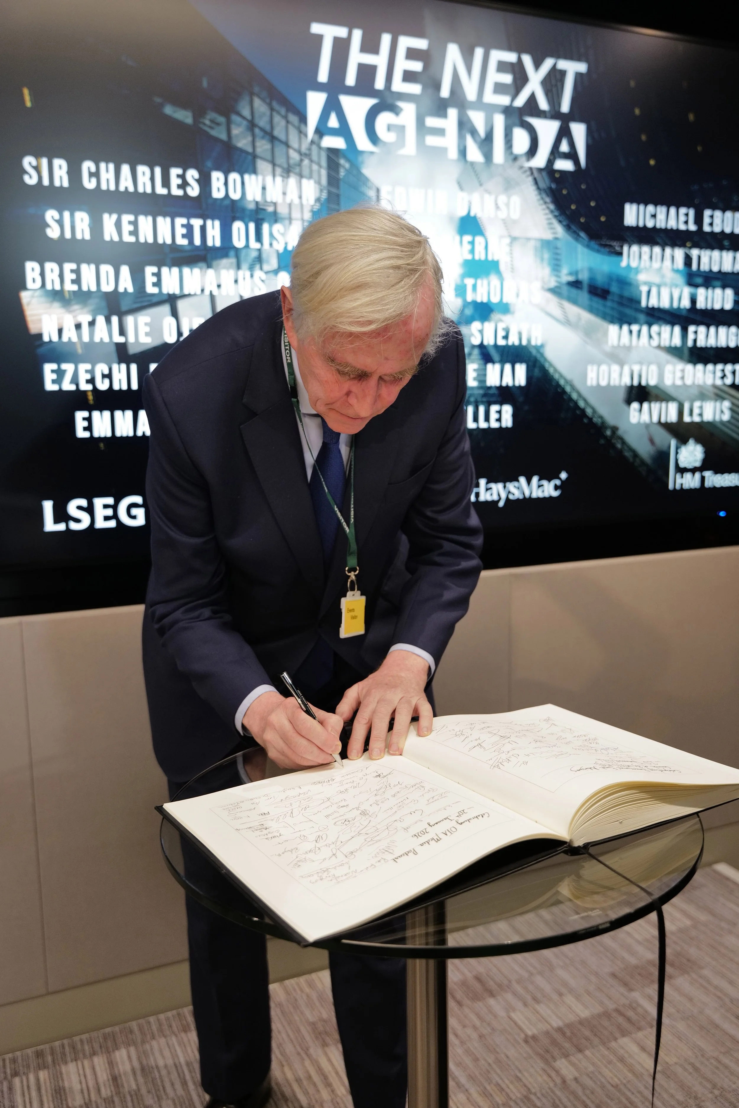 Man with light hair in a dark suit signing a large book on a glass table in front of a screen displaying 'The Next Agenda' and multiple names.