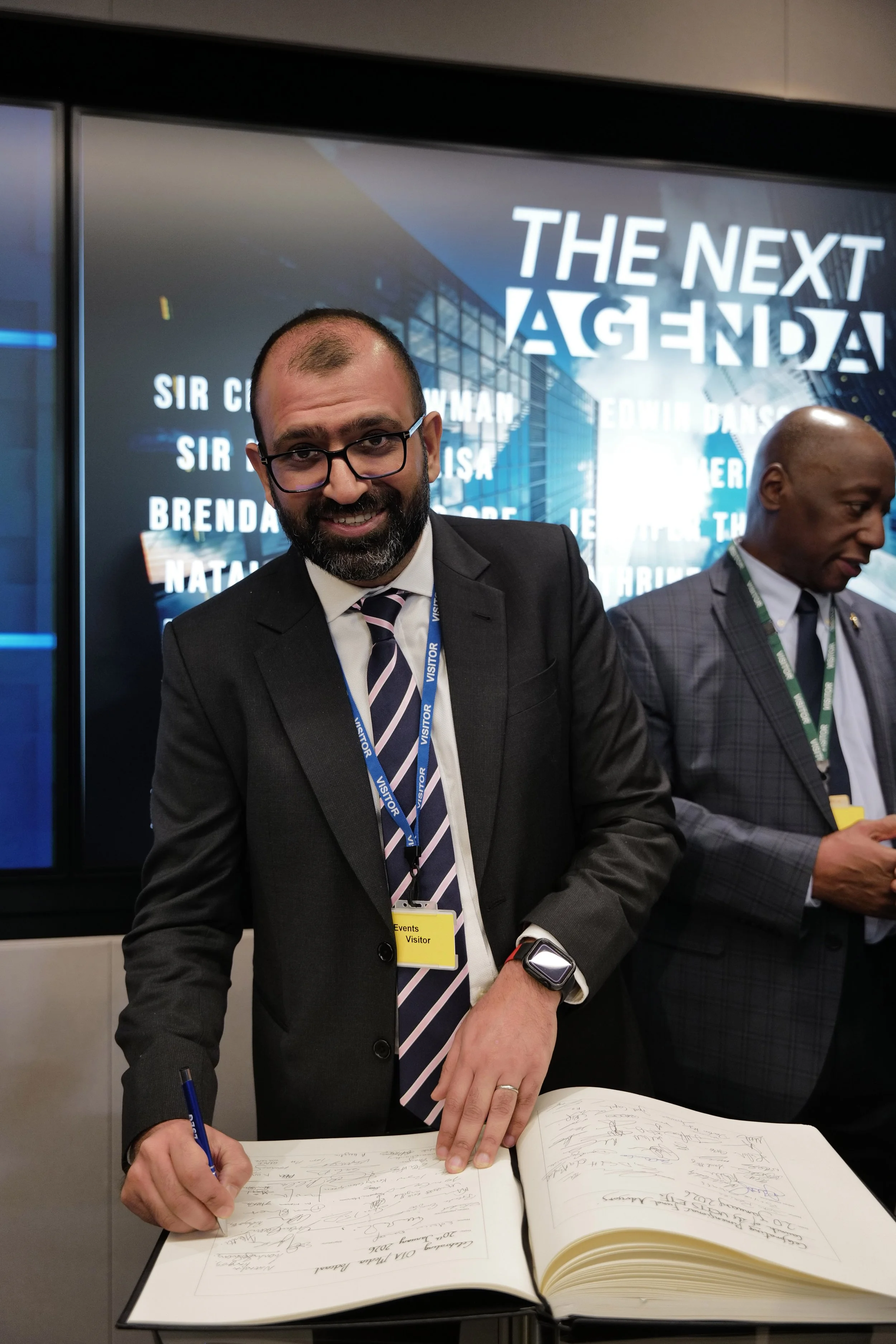 Man in glasses and suit signing a large book at an event with a visitor badge, another man in a suit is in the background.