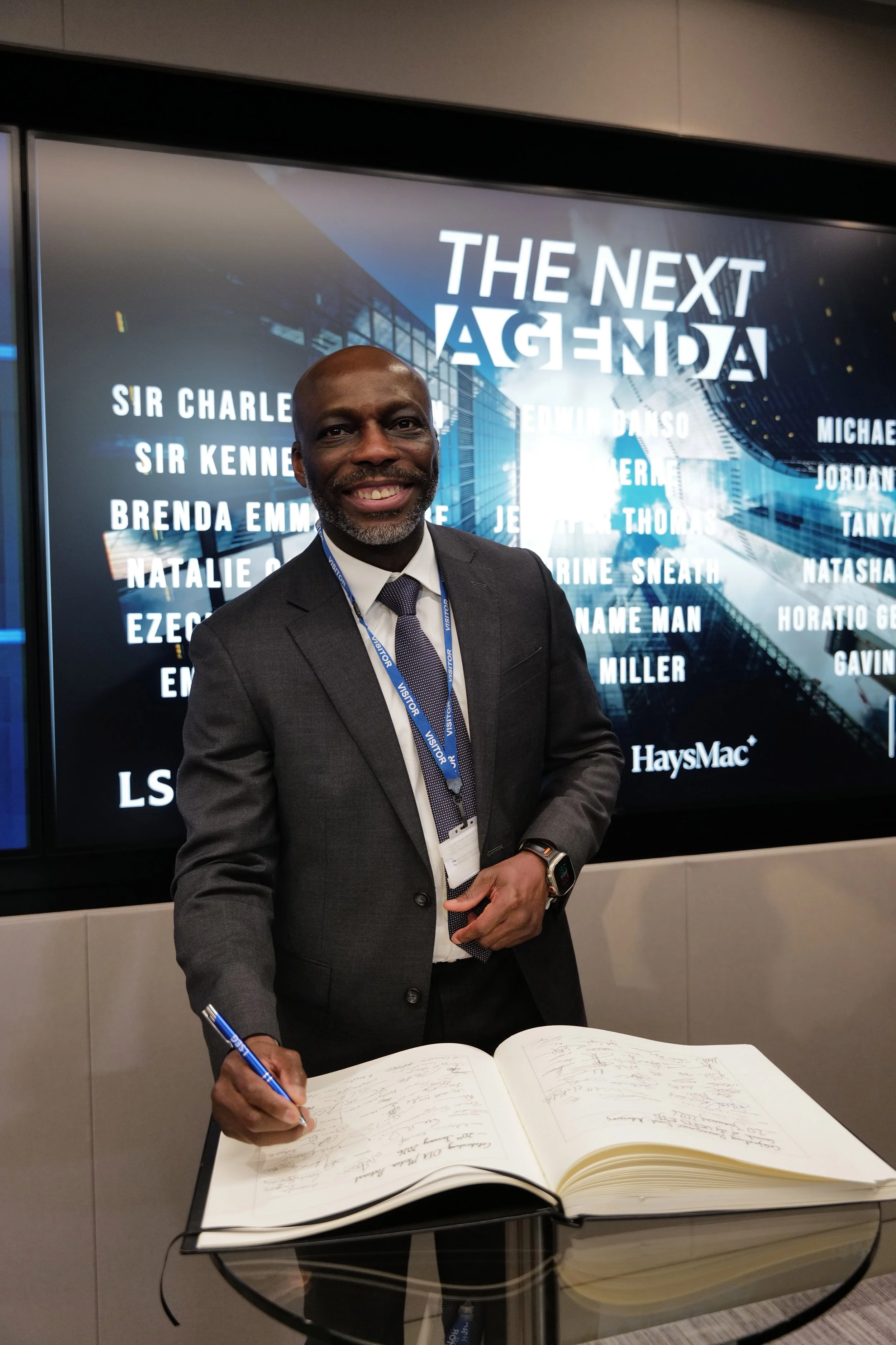 Smiling man in a suit signing a large book with a pen in front of a screen displaying 'THE NEXT AGENDA' and several names.