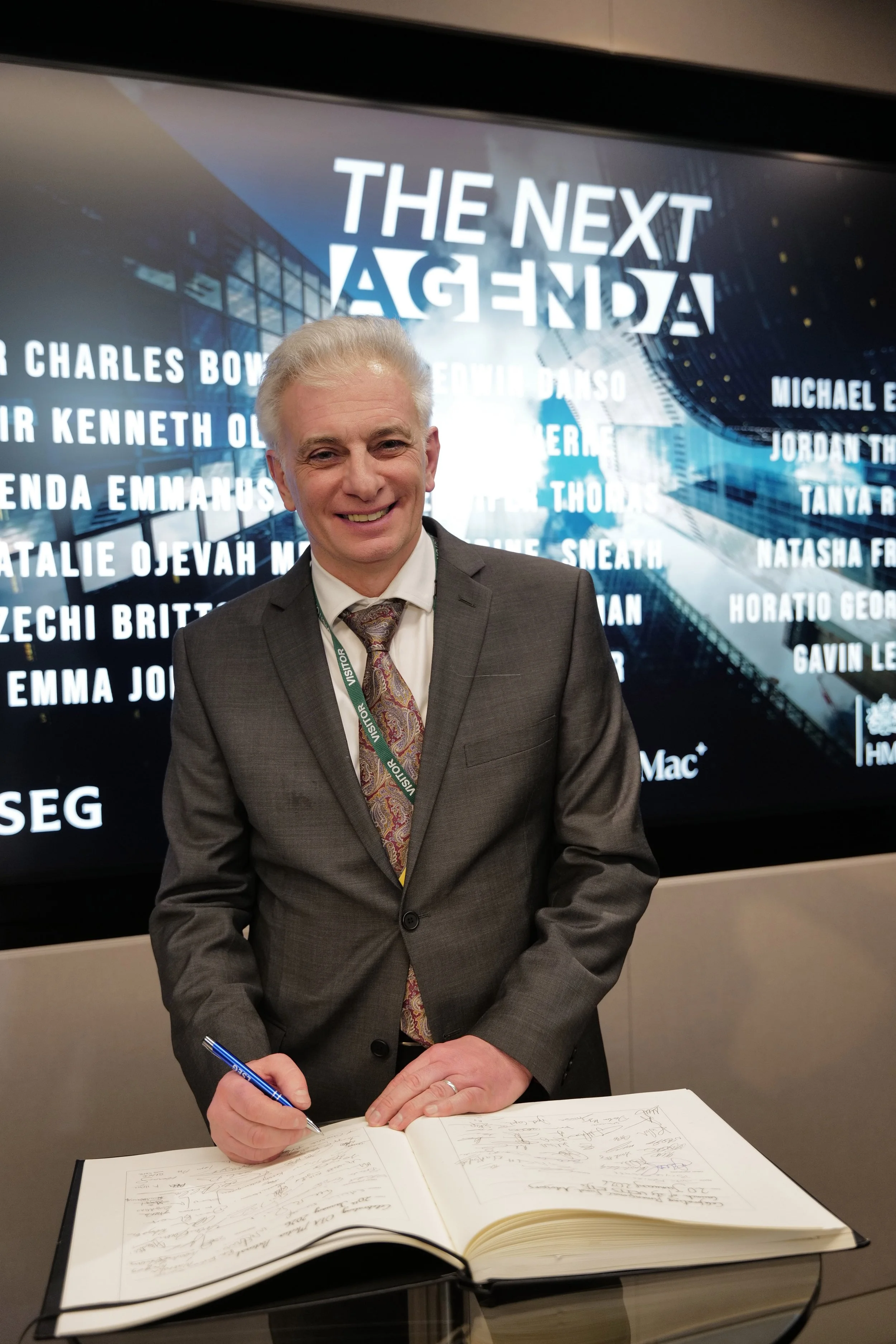 Man in gray suit signing a large book with a pen, standing in front of a screen displaying the text 'THE NEXT AGENDA'.