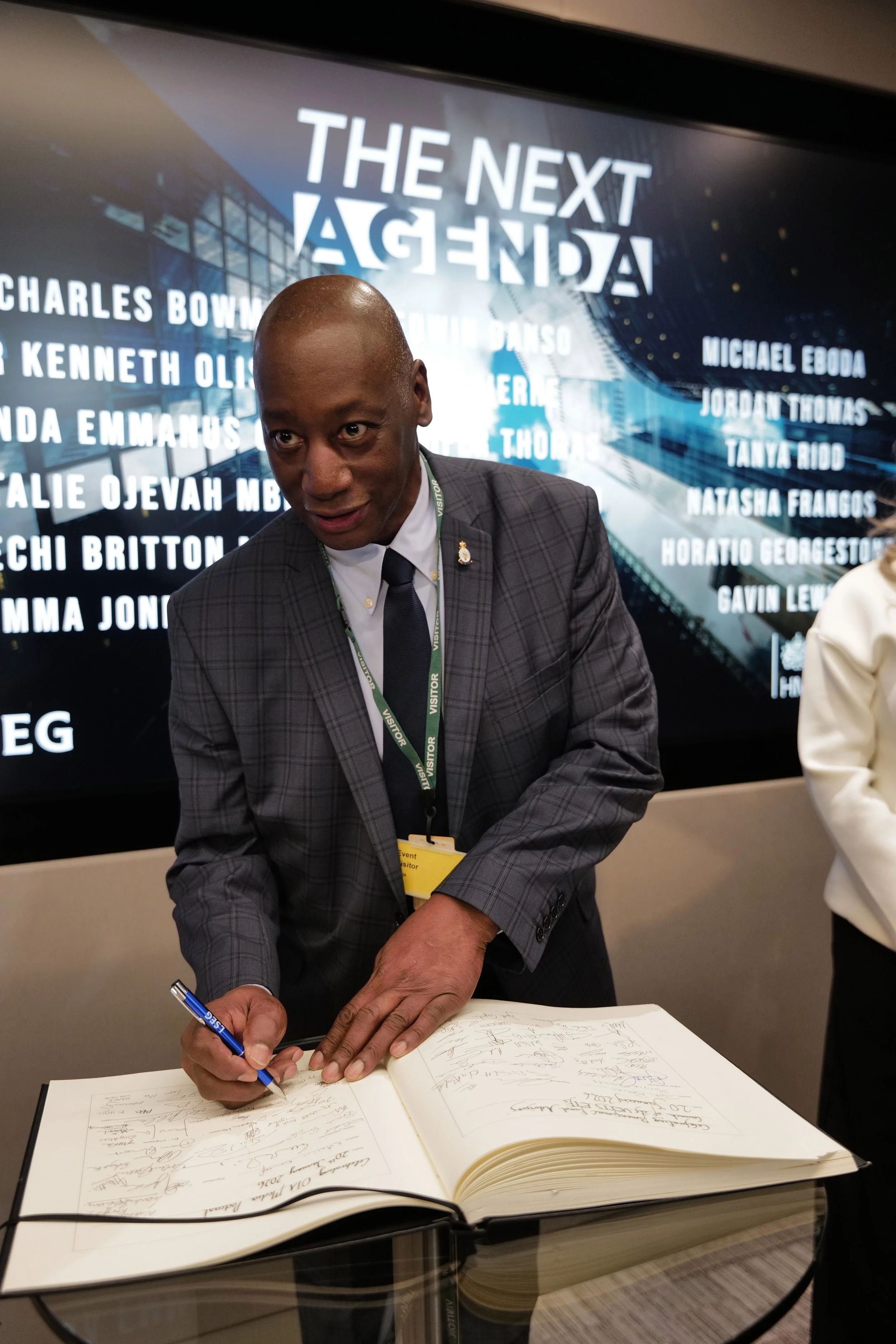 A man in a checkered suit jacket and dark tie signs a guest book at an event with a background screen displaying 'THE NEXT AGENDA' and multiple names.
