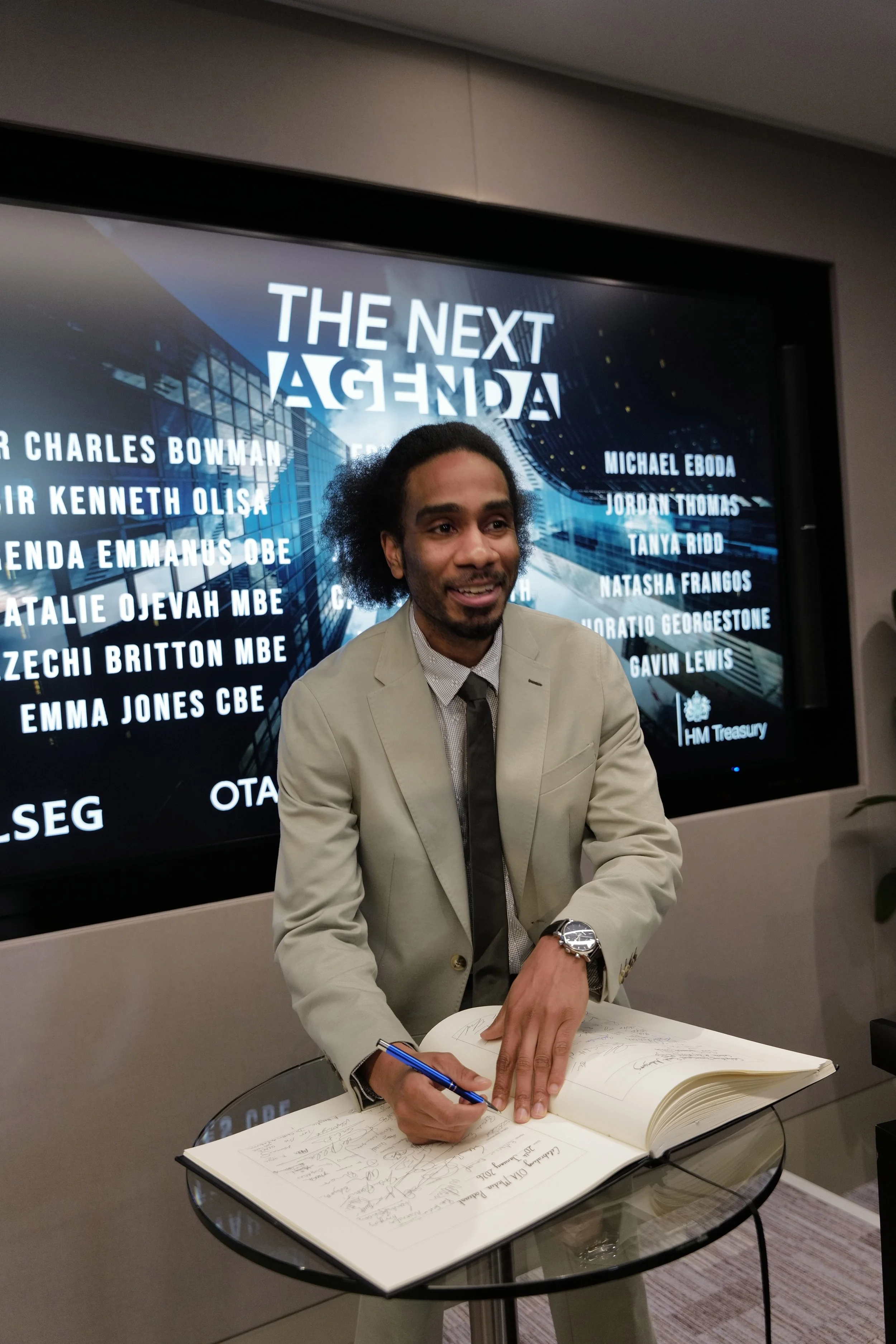 Man in beige suit signing a guestbook at an event with 'The Next Agenda' displayed on a screen behind him.
