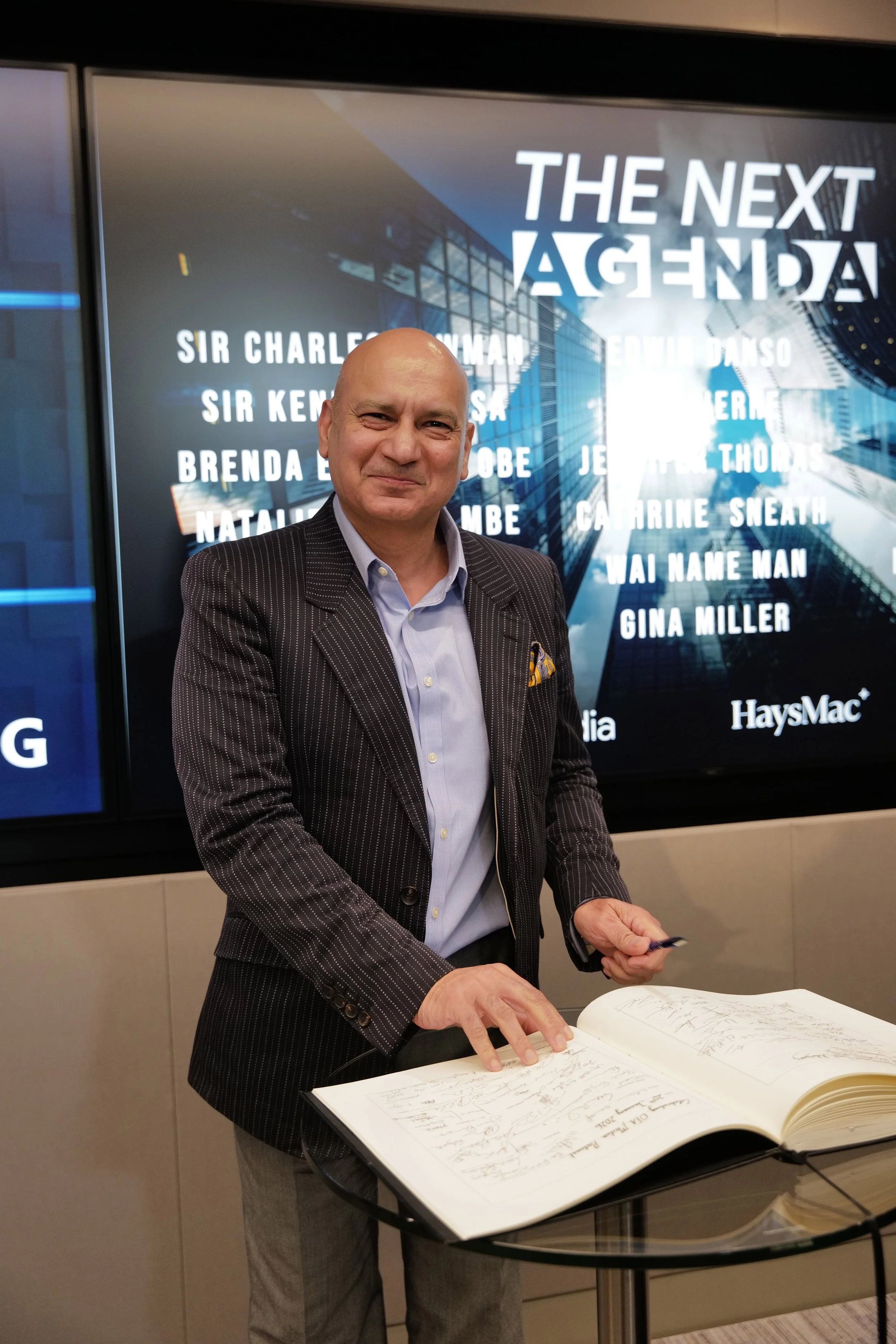 Smiling bald man in a pinstripe suit jacket signing a large book on a glass table with a backdrop reading THE NEXT AGENDA.
