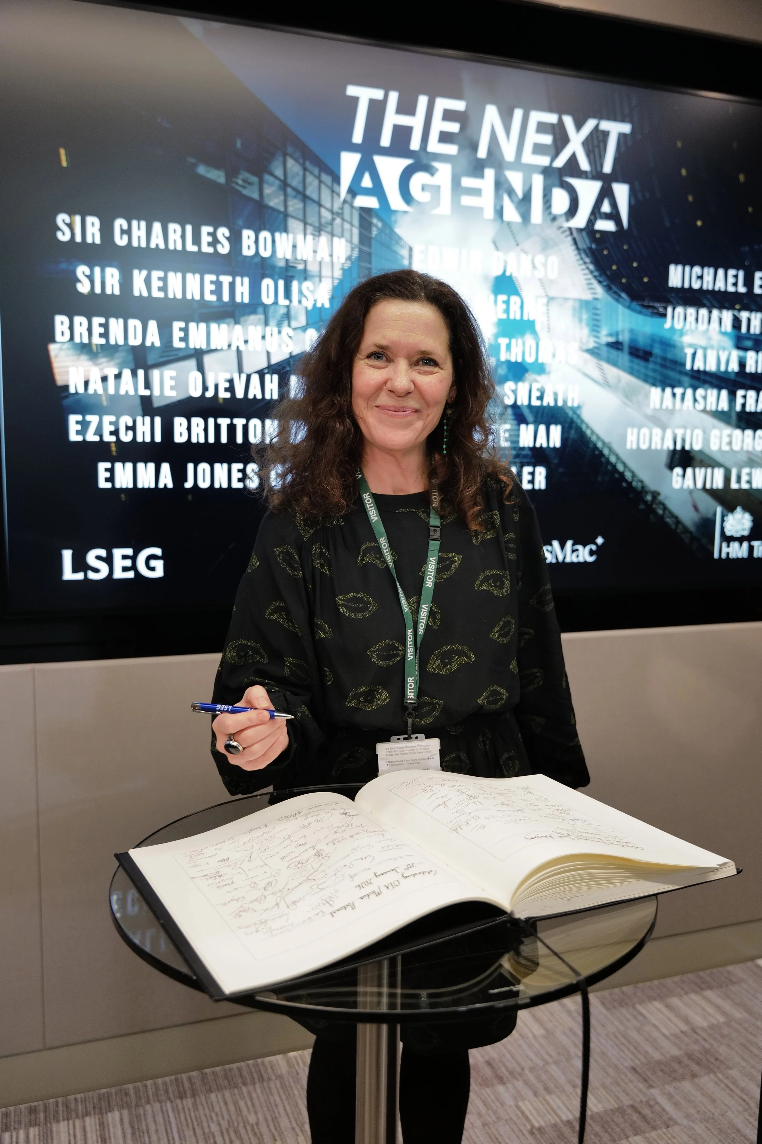 Smiling woman in a black dress standing behind a glass table signing a large guest book with a pen in front of a screen displaying 'The Next Agenda' and a list of names.
